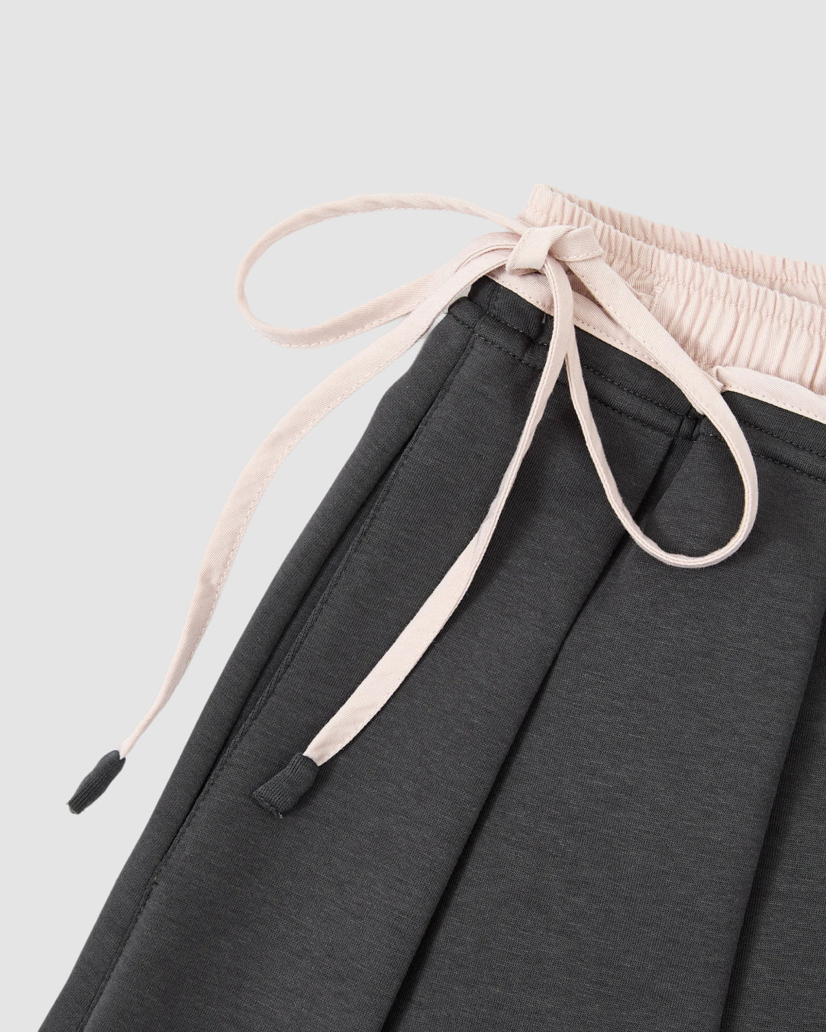 Close-up of the CHARCOAL PLEATED JERSEY MINI SKIRT by Sundae School, featuring a dark gray pleated design with a light pink elastic waistband and matching drawstring, set against a light gray background.