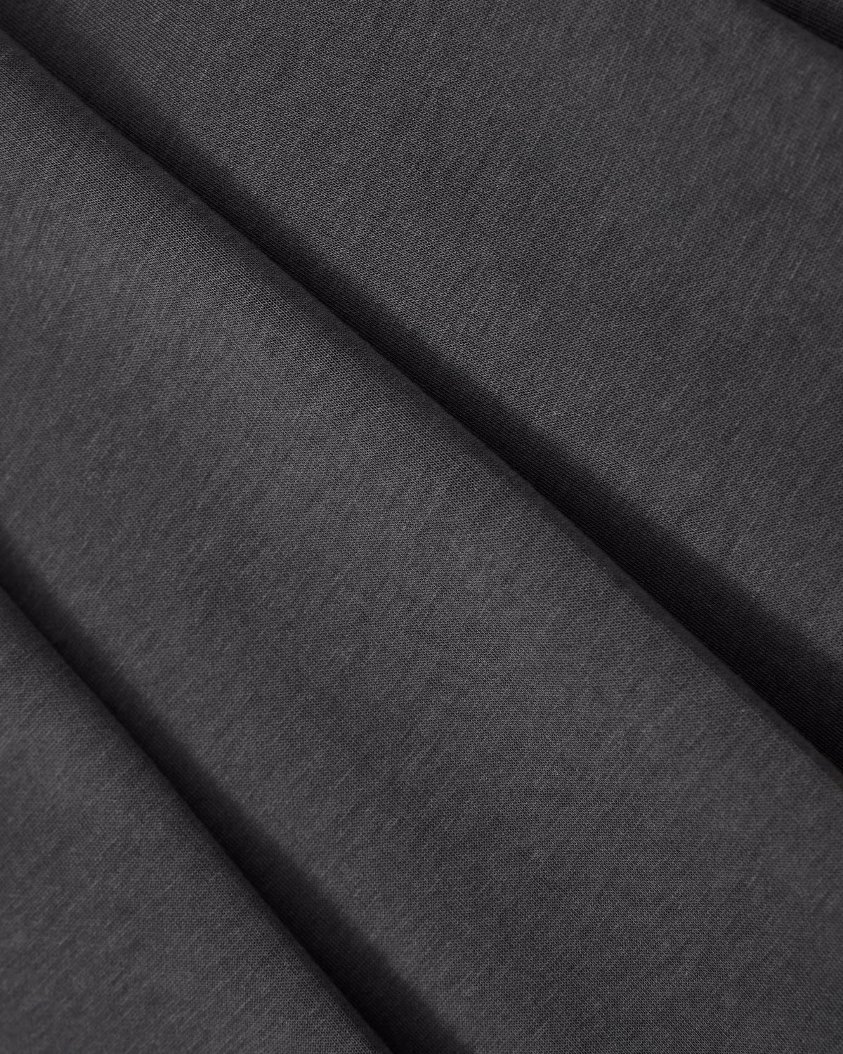A close-up of folded CHARCOAL PLEATED JERSEY MINI SKIRT by Sundae School, featuring dark gray, smooth, and soft cotton jersey fabric with subtle creases and shadows formed by the pleats.