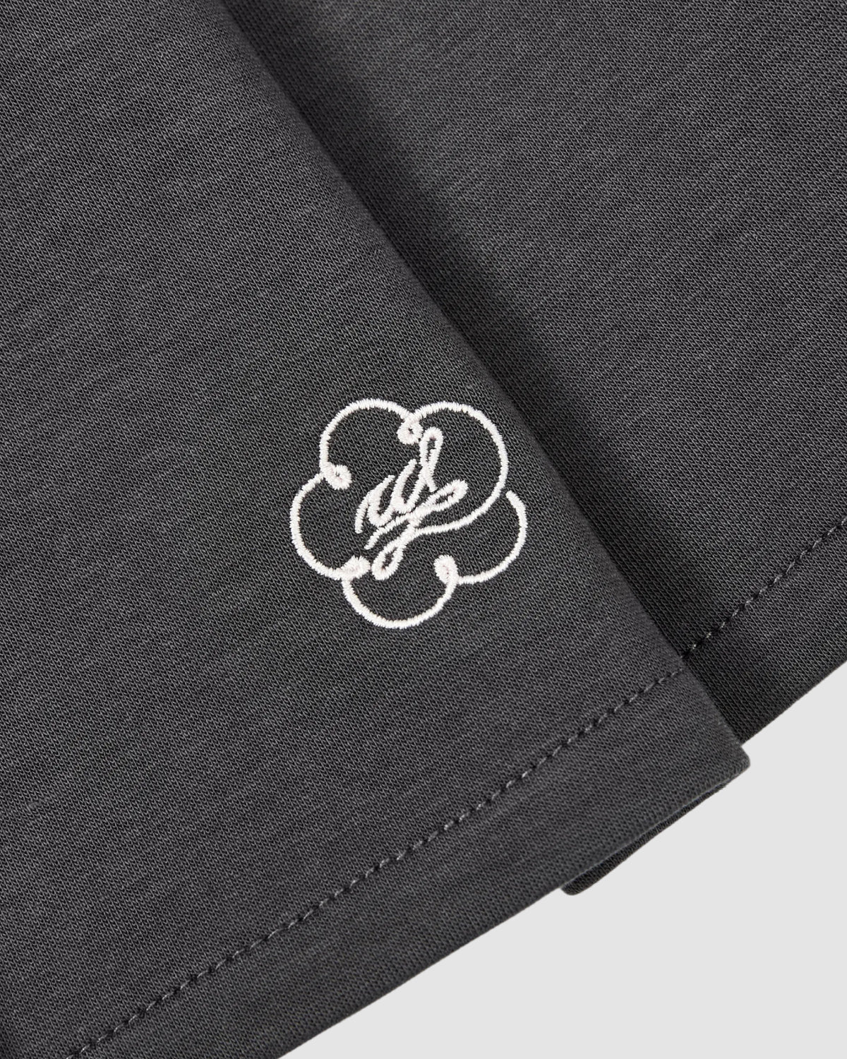 Close-up of the Sundae School CHARCOAL PLEATED JERSEY MINI SKIRT, featuring fine hem stitching and a white embroidered cloud logo with cursive initials on the edge.