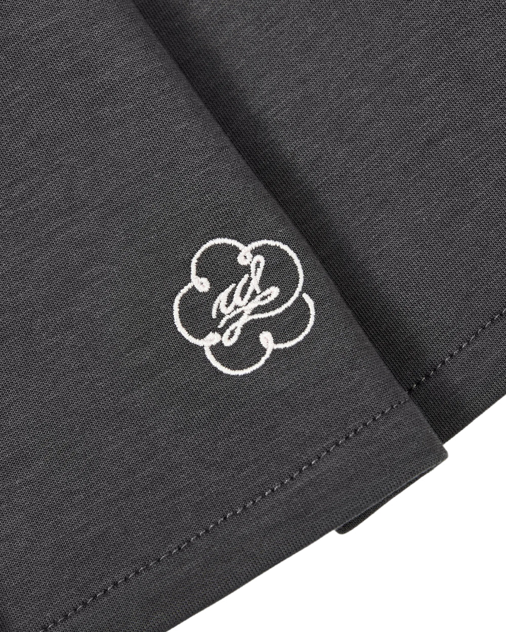 A close-up of the corner of the CHARCOAL PLEATED JERSEY MINI SKIRT by Sundae School, made from a dark gray cotton-blend jersey, featuring white stitching and an embroidered white cloud-like logo with cursive text.