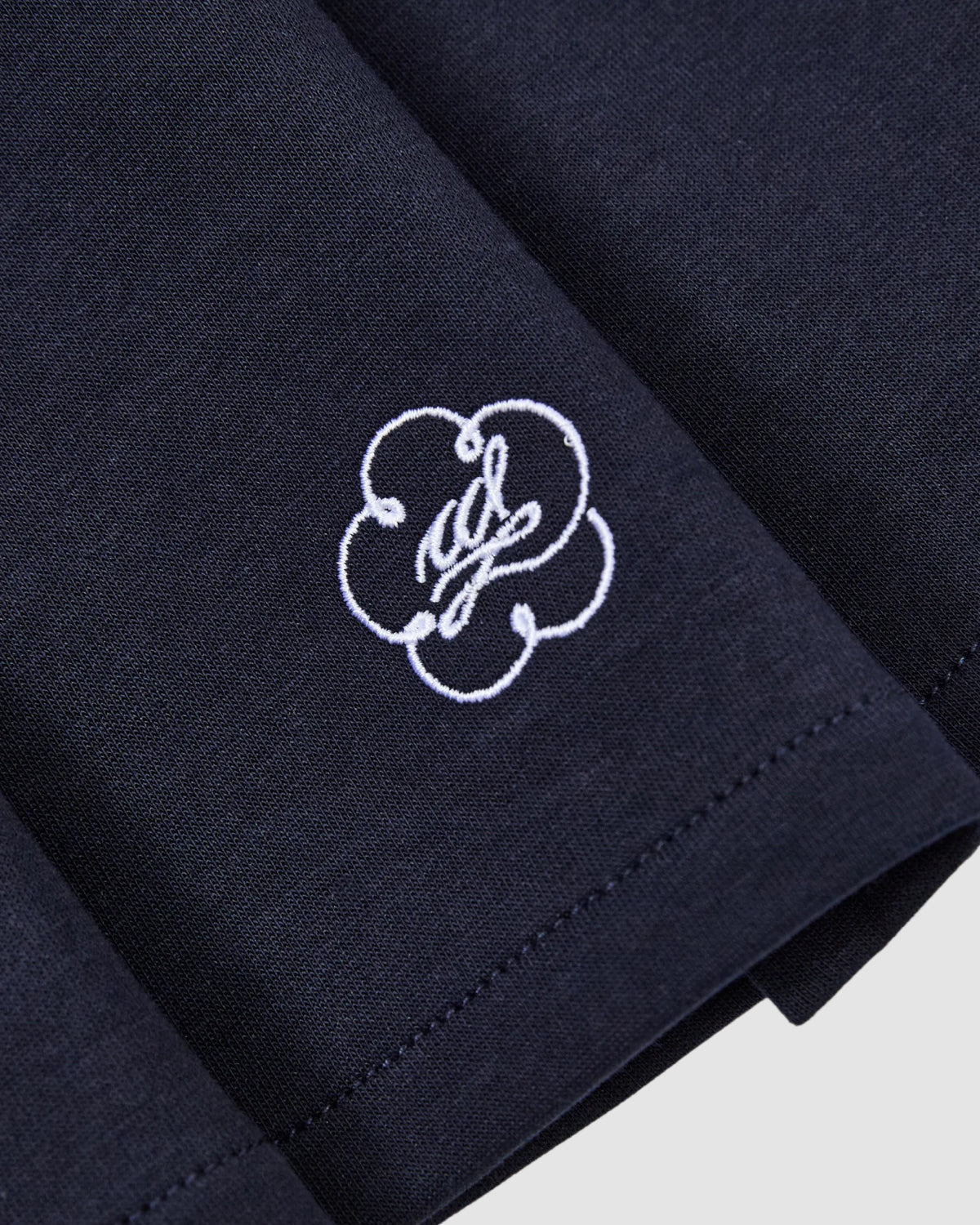 A close-up of the Sundae School NAVY PLEATED JERSEY MINI SKIRT shows neat stitching and a central white embroidered floral emblem with cursive lettering.