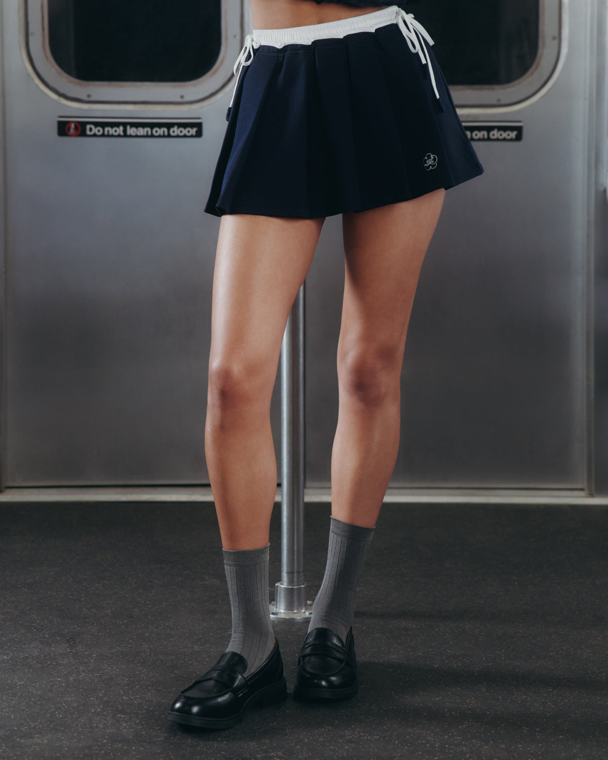 A person stands inside a subway train, wearing Sundae School's NAVY PLEATED JERSEY MINI SKIRT, paired with gray socks and black loafers; their upper body is out of frame.