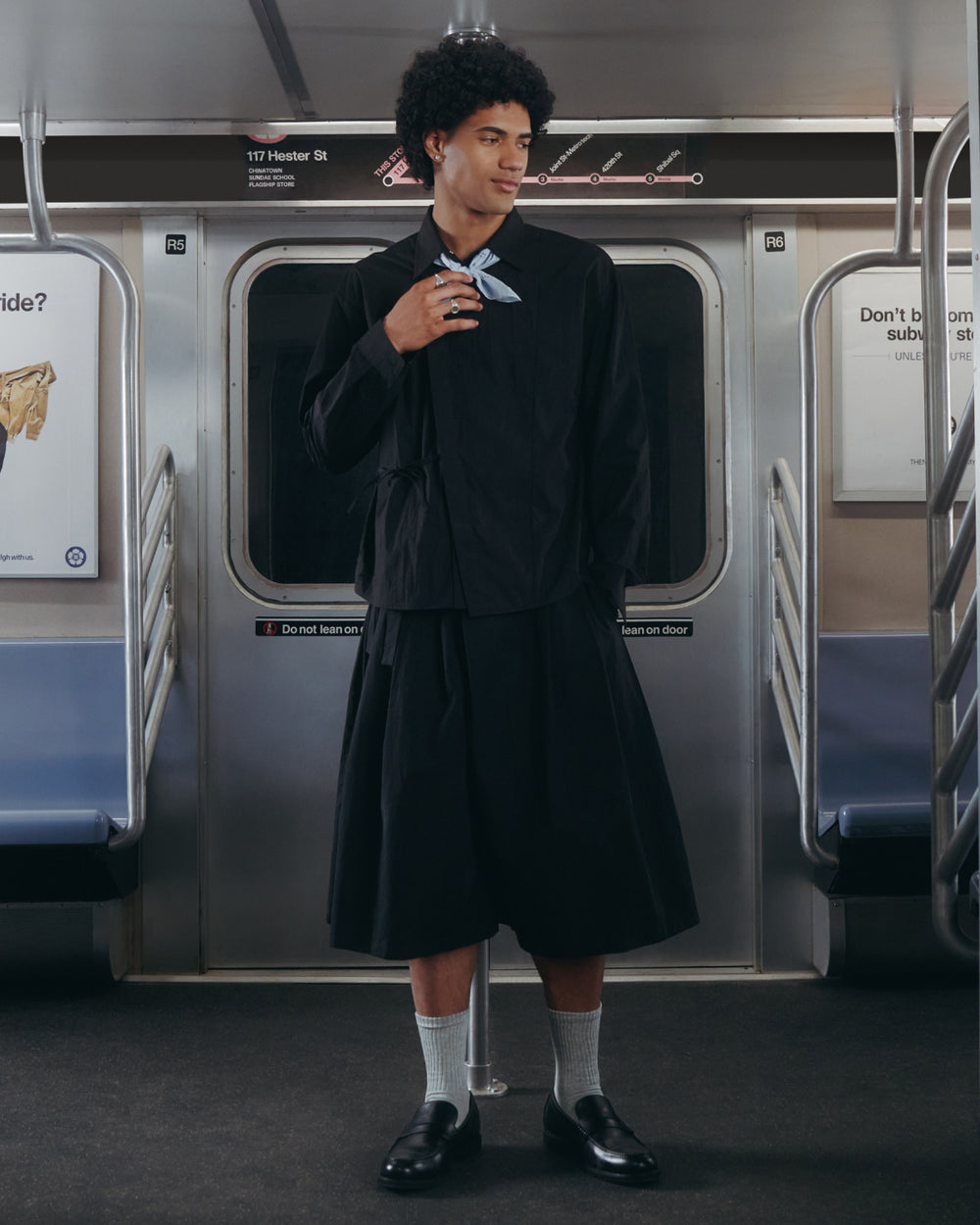 A person wearing the BLACK POPLIN TIE GORUM SHIRT by Sundae School, paired with a black skirt and dress shoes, stands in a subway car, looking to the side. The car has empty seats and signs on the walls.