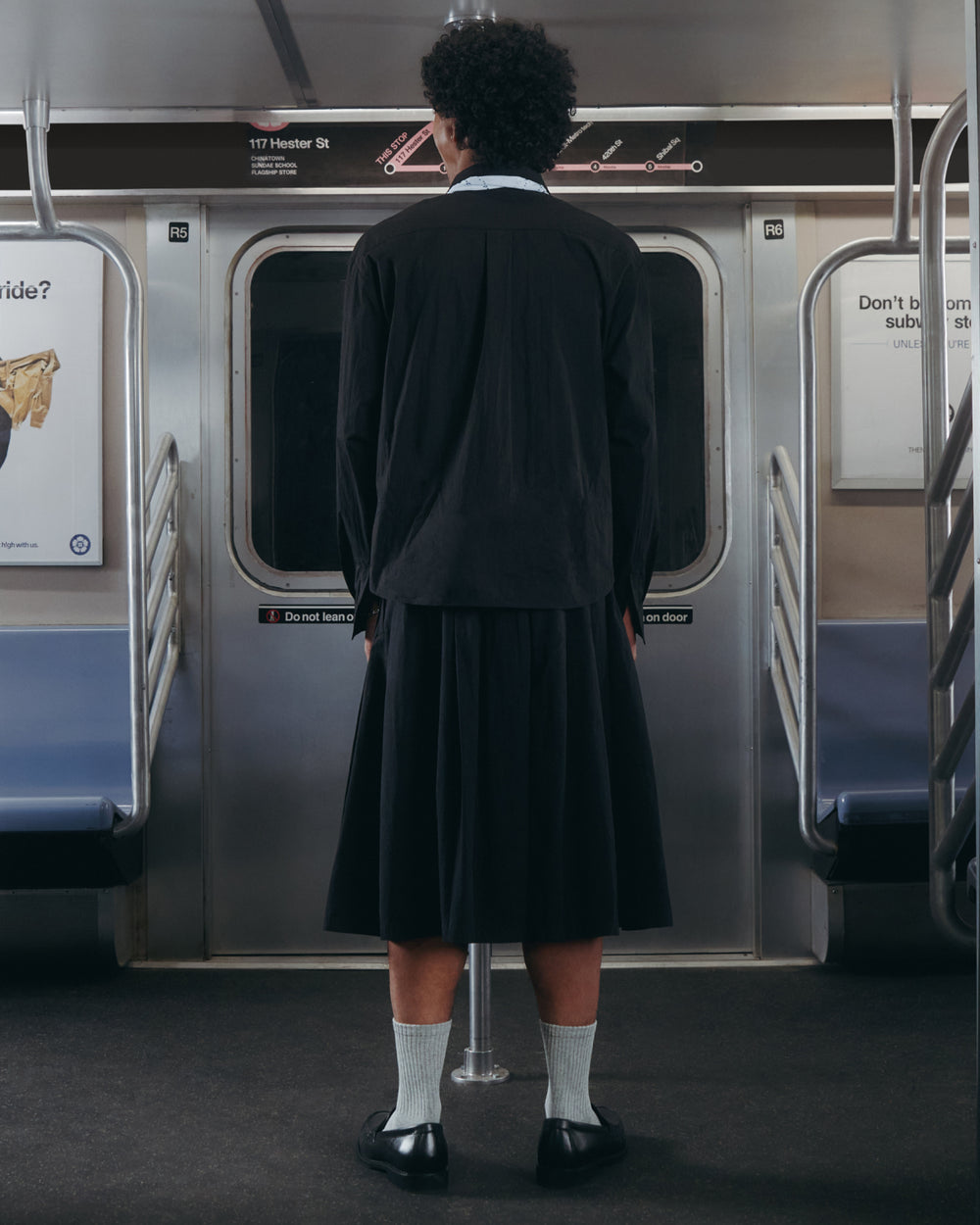 Wearing the BLACK POPLIN TIE GORUM SHIRT by Sundae School, a person with short curly hair, dressed in a black jacket, black skirt, white socks, and black shoes stands facing the doors inside an empty subway car.