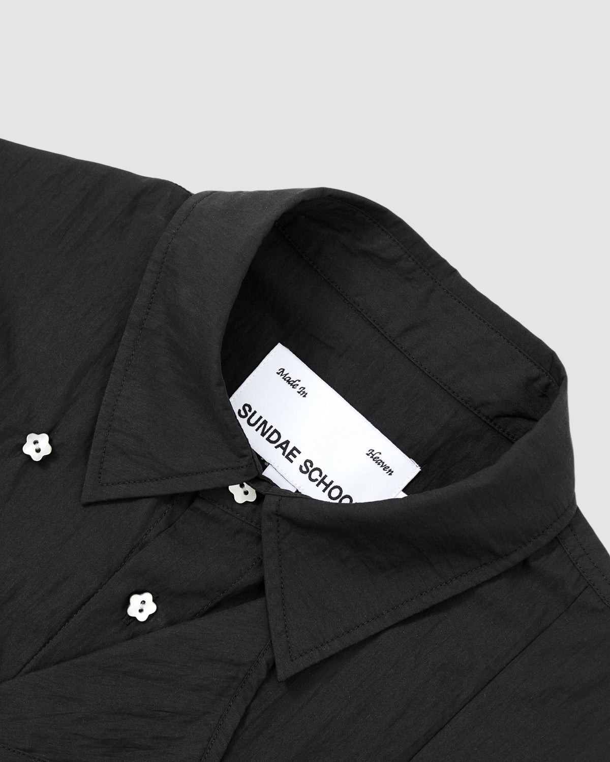 Close-up of the BLACK POPLIN TIE GORUM SHIRT by Sundae School, featuring white flower-shaped buttons and a visible inside label. The black collared shirt is set against a plain light gray background.