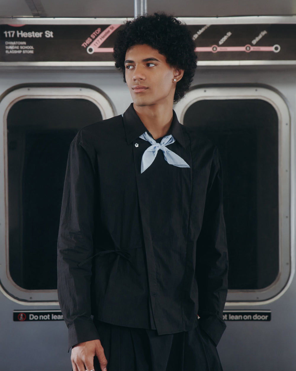 Wearing the BLACK POPLIN TIE GORUM SHIRT by Sundae School and a light blue scarf, a person stands in a subway train in front of a sign that reads "117 Hester St," glancing to the side as the closed train doors frame them.