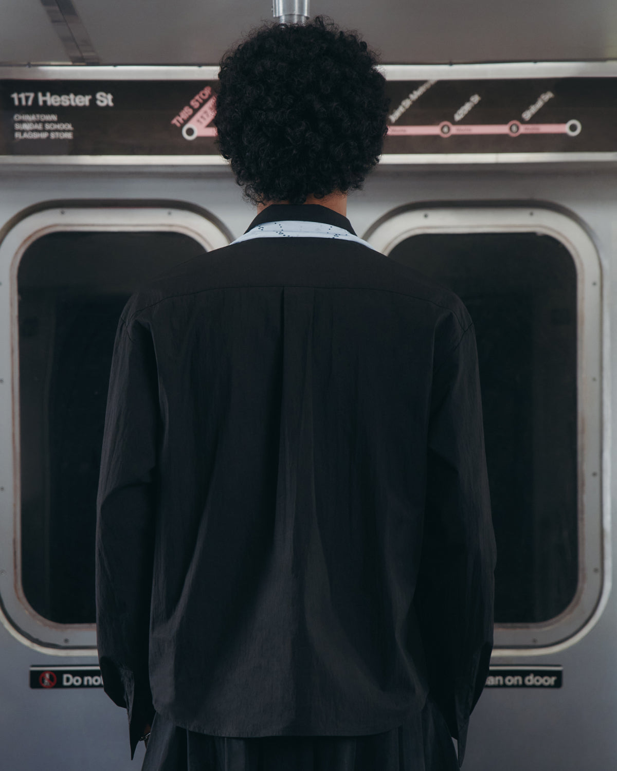 Wearing the Sundae School BLACK POPLIN TIE GORUM SHIRT, a person with curly hair stands inside a train at the 117 Hester St station, facing the subway doors with their back to the camera and the station name displayed above.