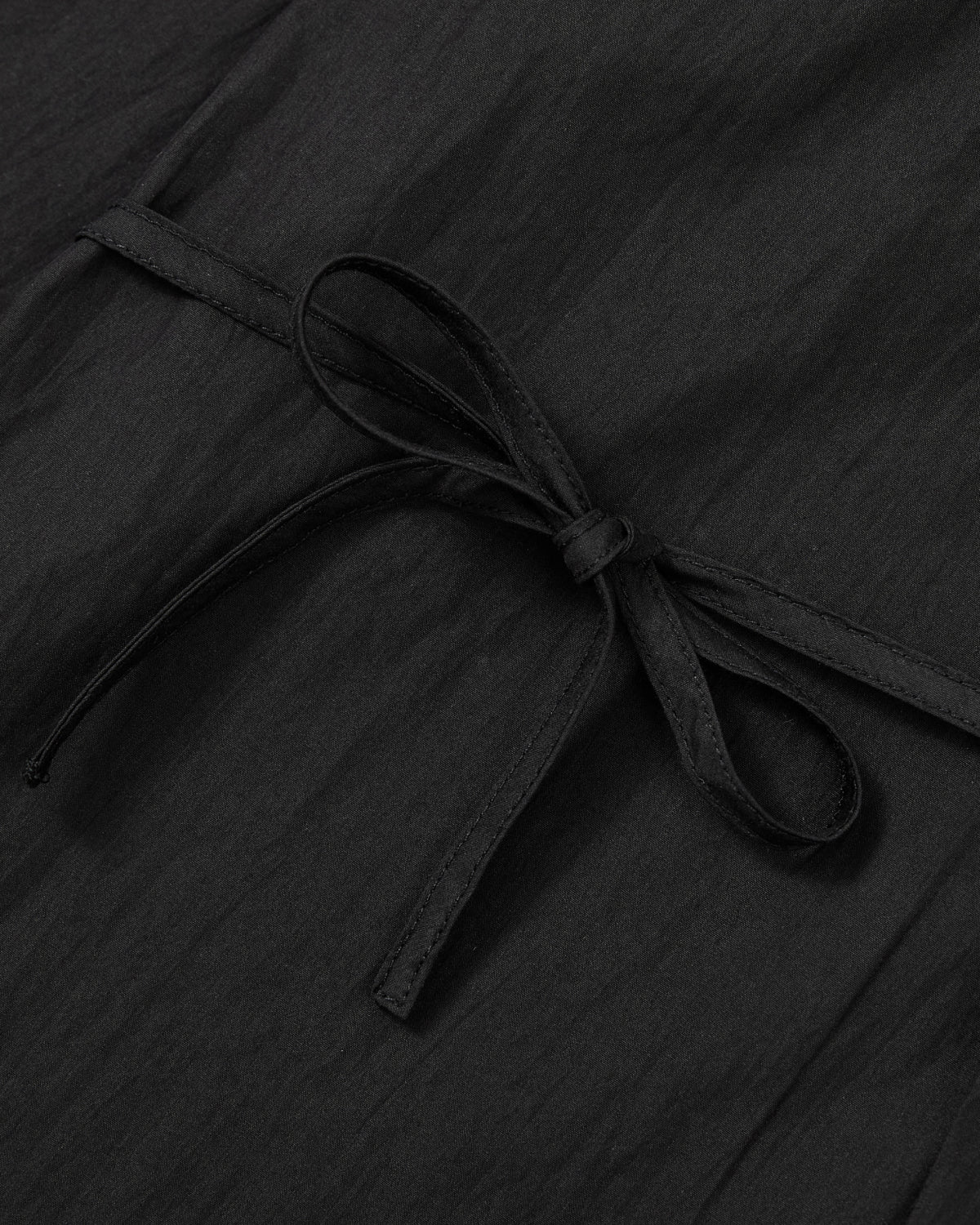 A close-up of the Sundae School BLACK POPLIN TIE GORUM SHIRT highlights its slightly crinkled poplin weave, with a neat bow knot formed from the shirt itself. The textured fabric and tailored unisex fit are clearly showcased in this detailed shot.