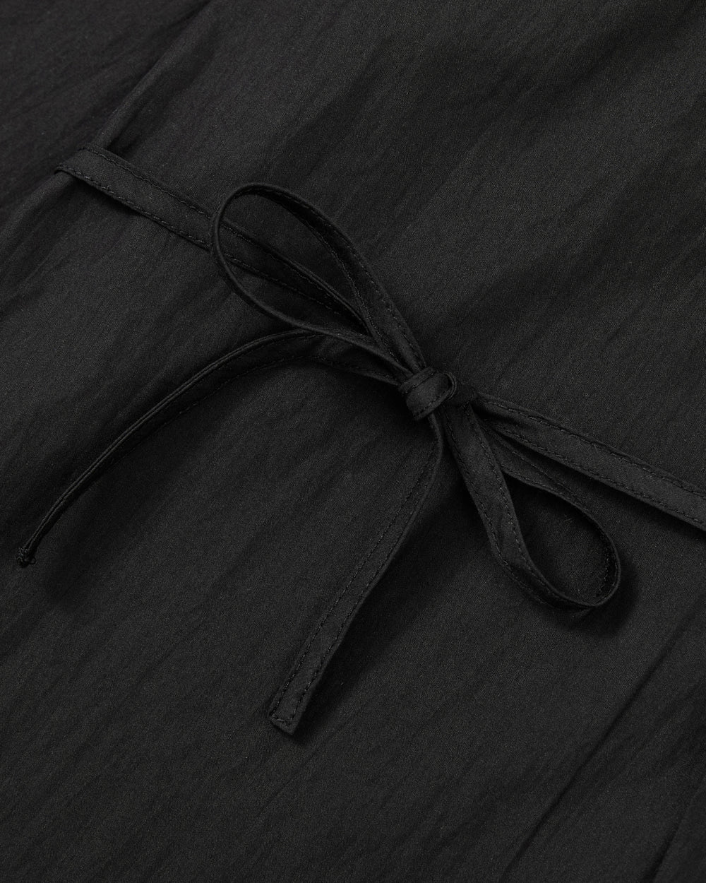 A close-up of the Sundae School BLACK POPLIN TIE GORUM SHIRT highlights its slightly crinkled poplin weave, with a neat bow knot formed from the shirt itself. The textured fabric and tailored unisex fit are clearly showcased in this detailed shot.