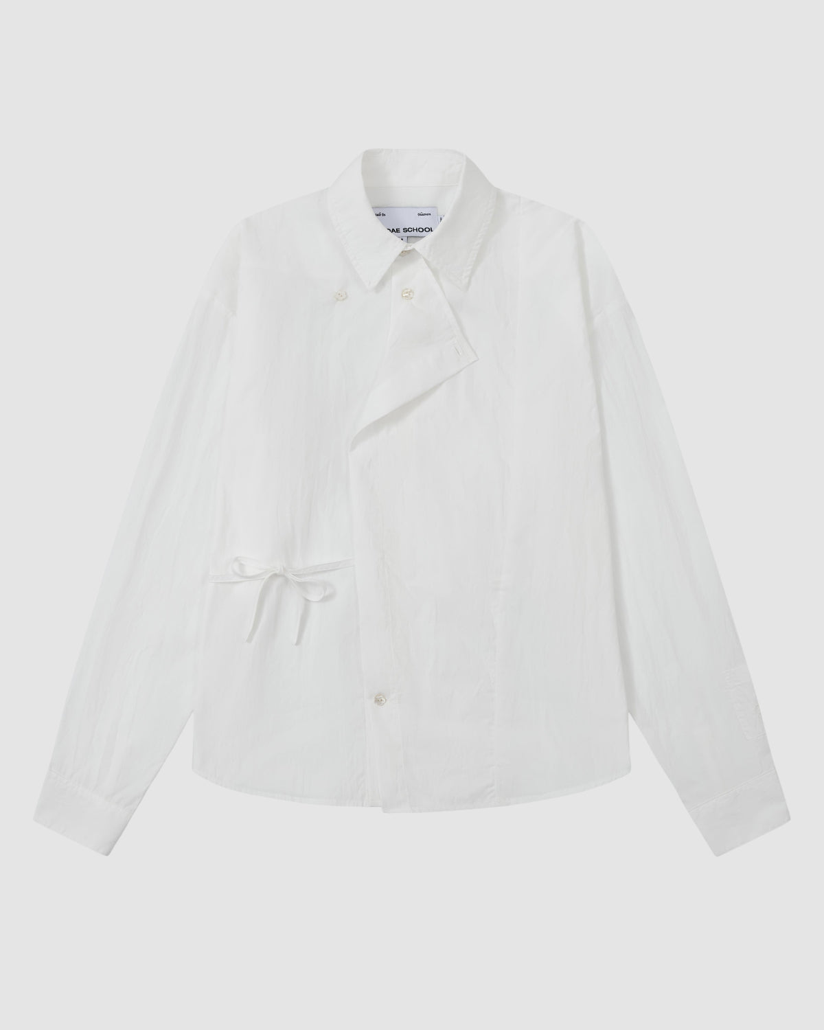 The WHITE POPLIN TIE GORUM SHIRT by Sundae School, a long-sleeve white button-up featuring an asymmetric front and side tie detail, is laid flat on a light background.