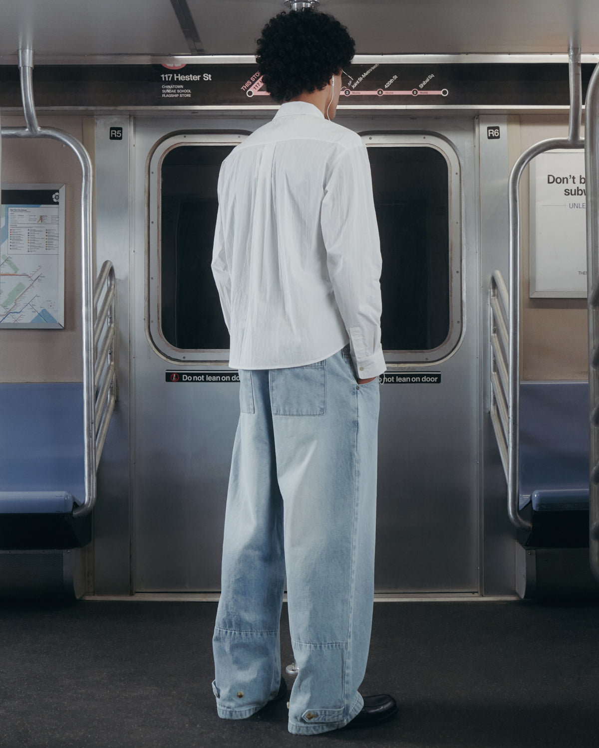 Wearing the WHITE POPLIN TIE GORUM SHIRT by Sundae School and baggy light blue jeans, a person with curly hair stands facing subway doors inside a train car.
