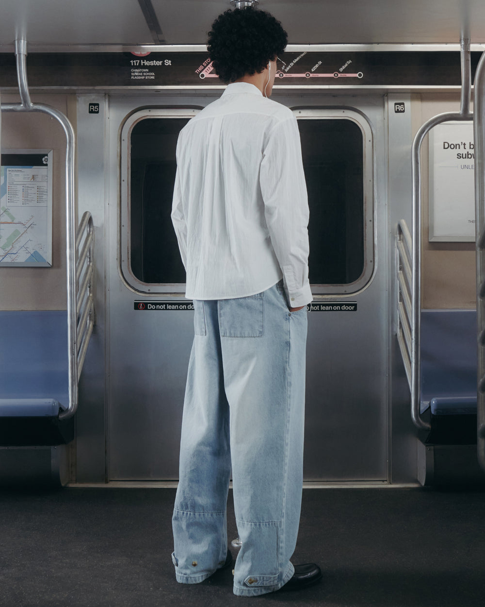 Wearing the WHITE POPLIN TIE GORUM SHIRT by Sundae School and baggy light blue jeans, a person with curly hair stands facing subway doors inside a train car.
