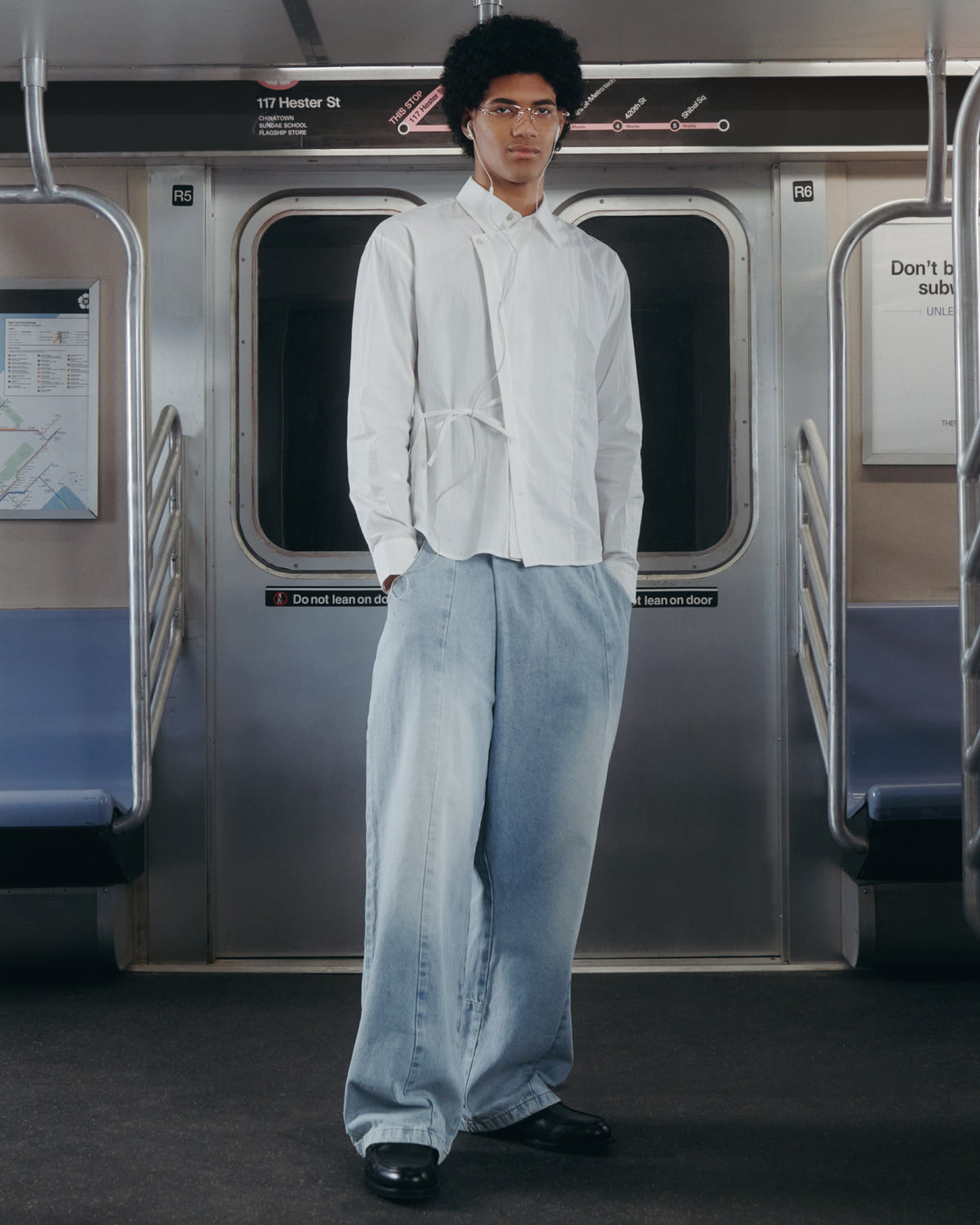A person wearing the WHITE POPLIN TIE GORUM SHIRT by Sundae School and wide, light blue jeans stands in the middle of an empty subway car, facing the camera with hands in their pockets. Subway signs and metal doors appear in the background.