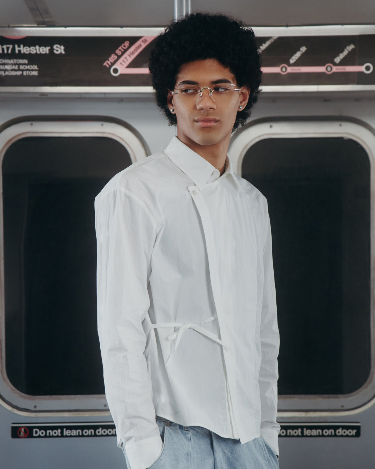 Wearing the WHITE POPLIN TIE GORUM SHIRT by Sundae School, a person with curly hair and glasses stands in a subway train near a Hester St. station sign, gazing to the side.