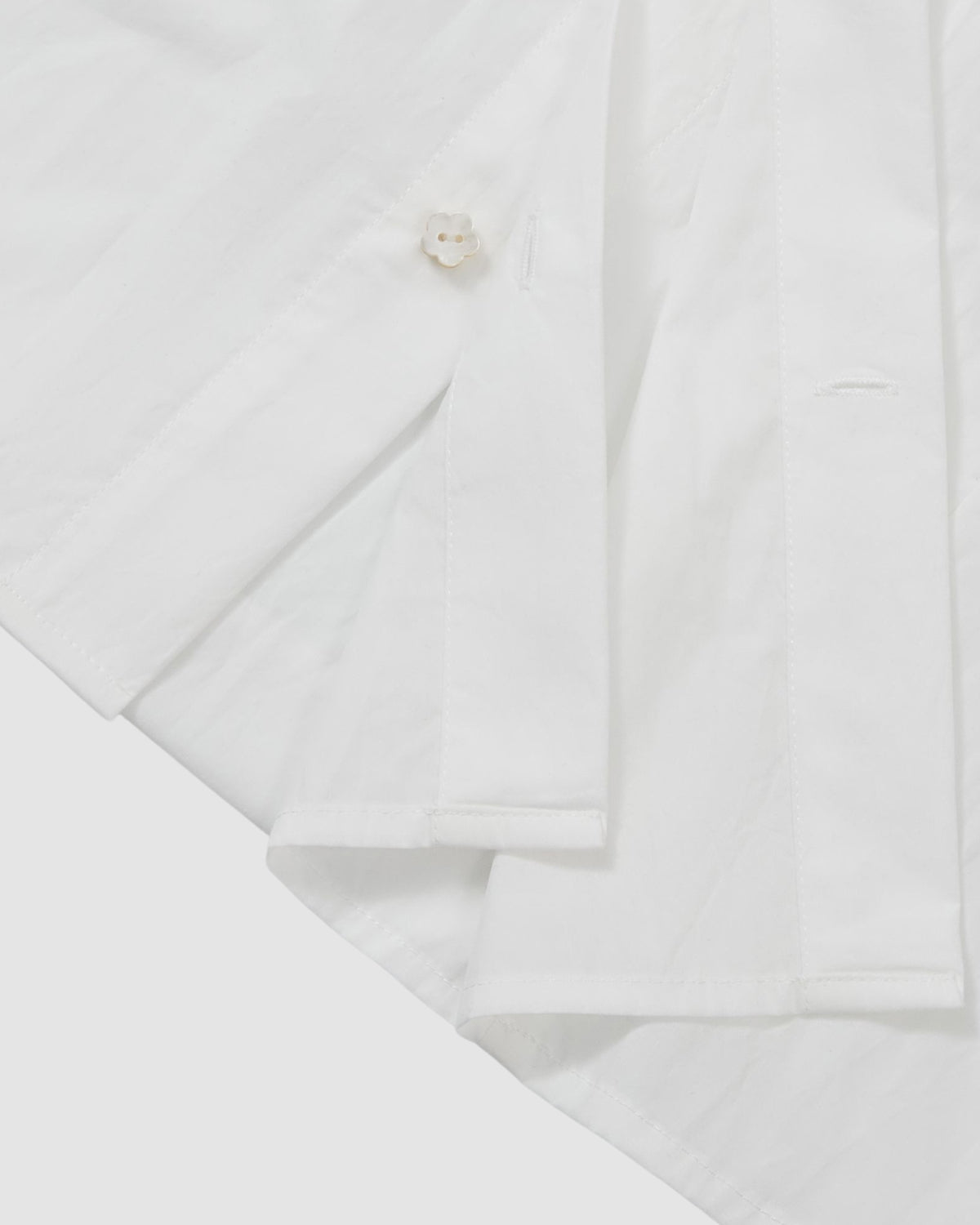 Close-up of the WHITE POPLIN TIE GORUM SHIRT by Sundae School, highlighting the hem, a button, and part of the sleeve against a white background.