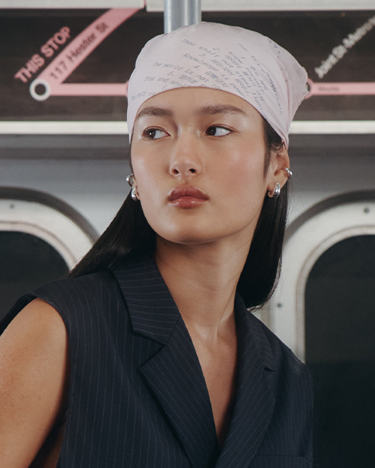 A woman in the Sundae School BABY PINK 10 COMMANDMENTS ORGANZA BANDANA, dark pinstripe vest, and silver ear cuffs looks to the side on public transportation with a subway map in the background.