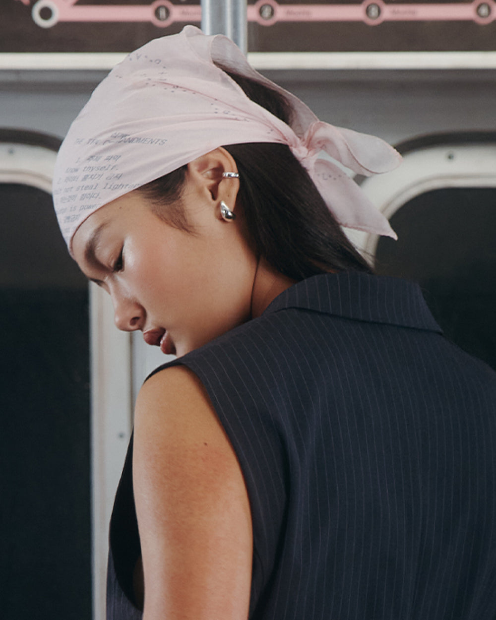 A woman with long dark hair wears the Sundae School BABY PINK 10 COMMANDMENTS ORGANZA BANDANA and a sleeveless navy pinstripe blazer, looking down with her head turned to the side. Two silver earrings adorn her ear.