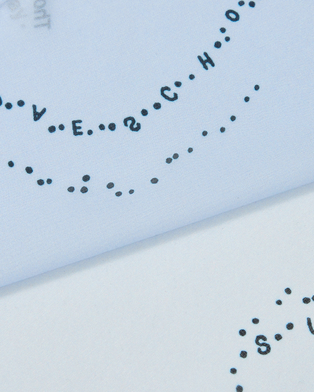 Close-up of a page with black ink letters and dots arranged in a curved line, partially visible on light blue paper. The lettering spells out SCHOOL and other words, evoking the bold printed texture found on the Sundae School SKY BLUE 10 COMMANDMENTS ORGANZA BANDANA.