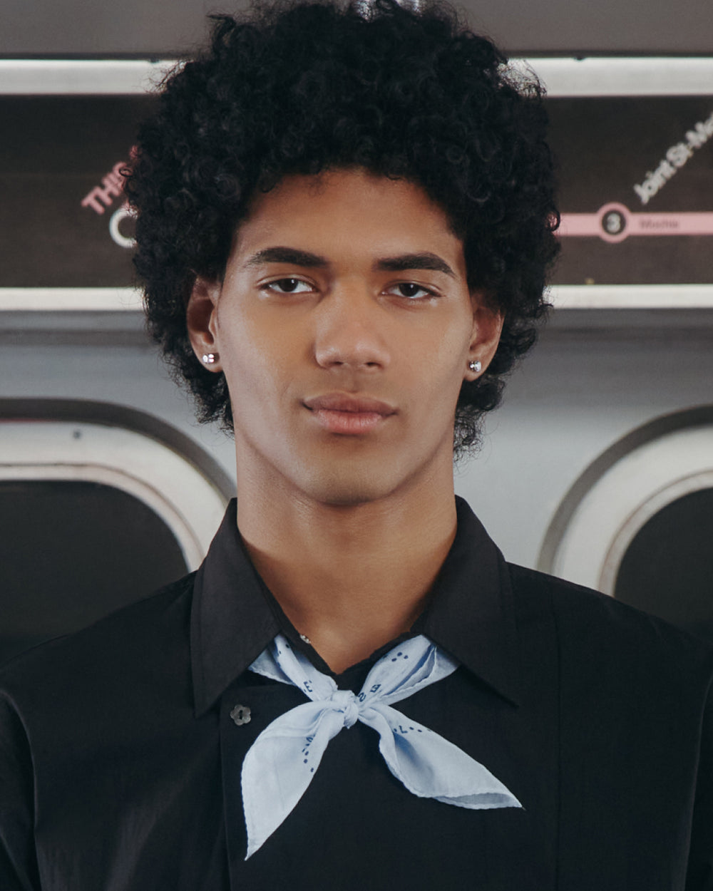 A young person with curly black hair, silver stud earrings, and a black shirt wears the Sundae School SKY BLUE 10 COMMANDMENTS ORGANZA BANDANA styled as a neck bandana while standing in front of a subway train interior.