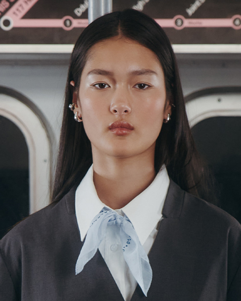 A person with long dark hair, in a gray jacket, white collared shirt, and Sundae School’s SKY BLUE 10 COMMANDMENTS ORGANZA BANDANA, stands inside a subway train with a transit map visible in the background.