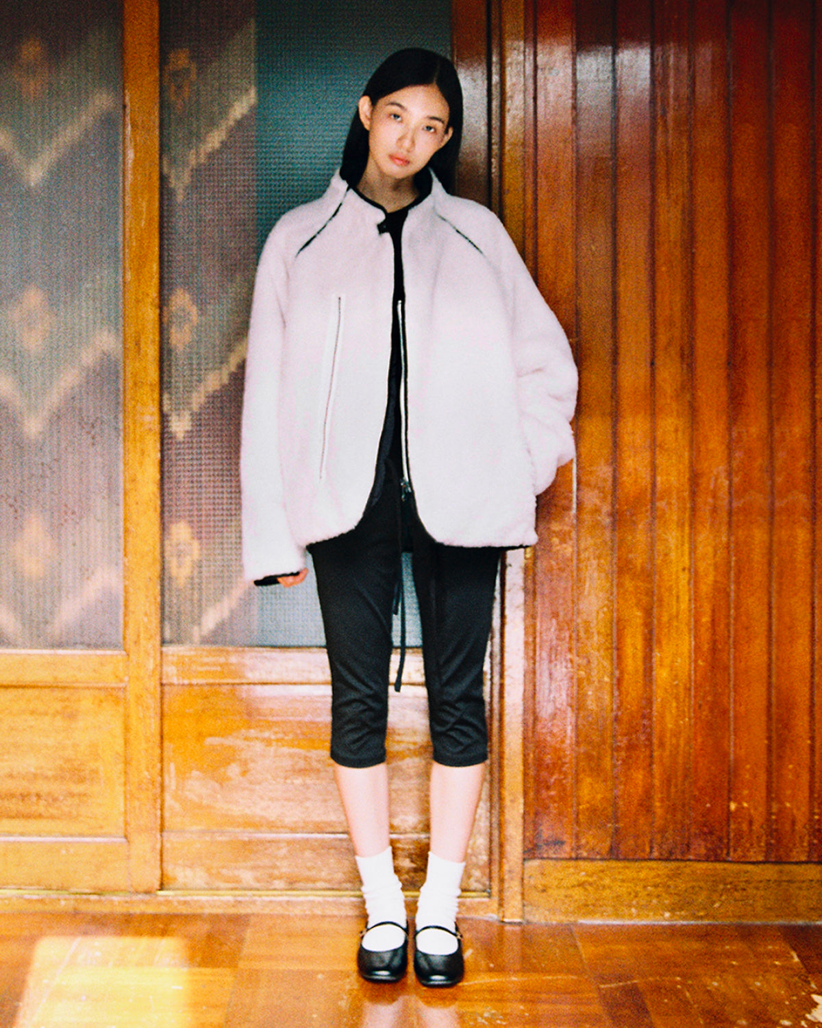 A woman stands indoors against wooden paneling, wearing Sundae School’s BLACK IVORY REVERSIBLE FLOWER QUILTED JEOGORI FLEECE, black cropped pants, white socks, and black Mary Jane shoes. She looks at the camera with a neutral expression.