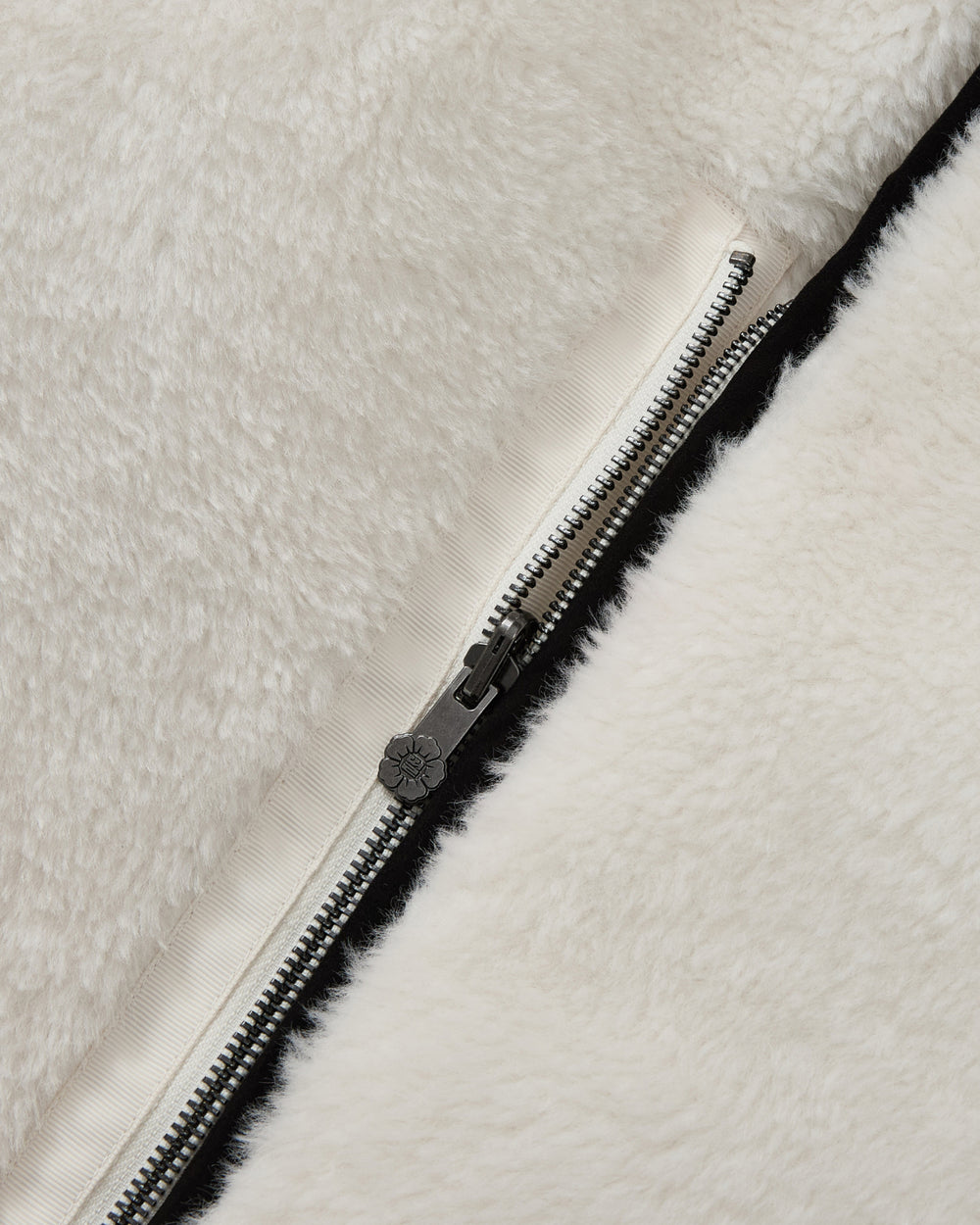Close-up of a silver zipper on the soft, white faux-sherpa fabric of the BLACK IVORY REVERSIBLE FLOWER QUILTED JEOGORI FLEECE by Sundae School, partially unzipped to reveal a beige inner lining.
