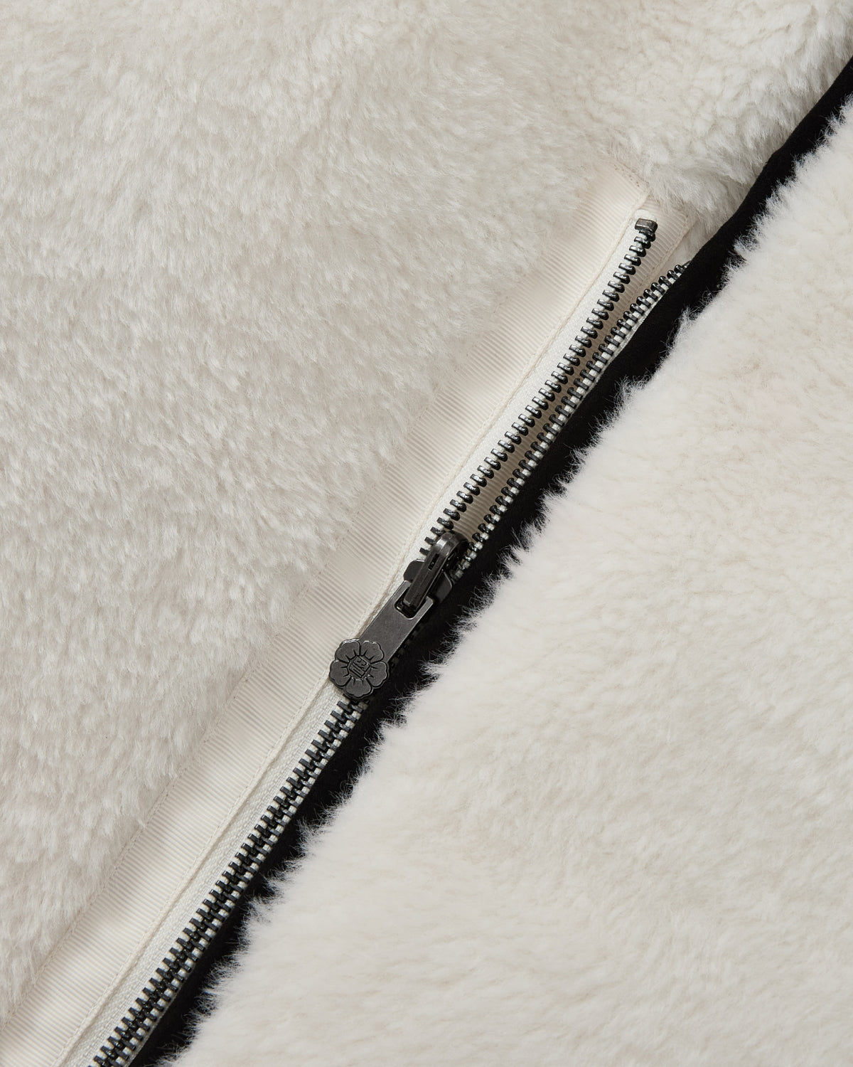 Close-up of a silver zipper on the soft, white faux-sherpa fabric of the BLACK IVORY REVERSIBLE FLOWER QUILTED JEOGORI FLEECE by Sundae School, partially unzipped to reveal a beige inner lining.