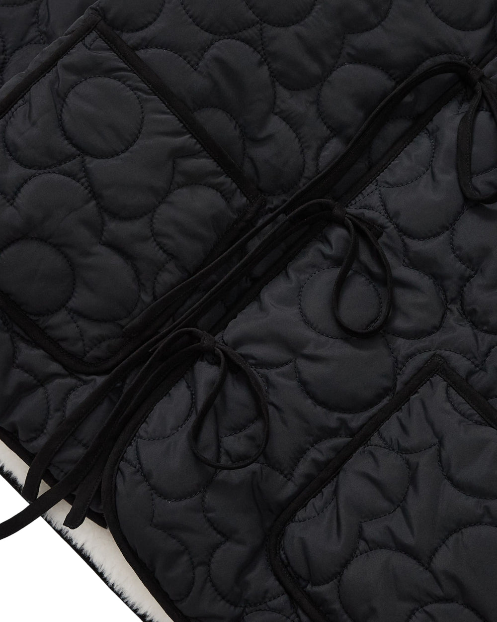 Close-up of the BLACK IVORY REVERSIBLE FLOWER QUILTED JEOGORI FLEECE by Sundae School, featuring black quilted fabric with circular stitching patterns, two spacious pockets, and long fabric ties. The visible texture and stitching highlight the coat’s hanbok-inspired design, emphasizing the material’s softness and thickness.