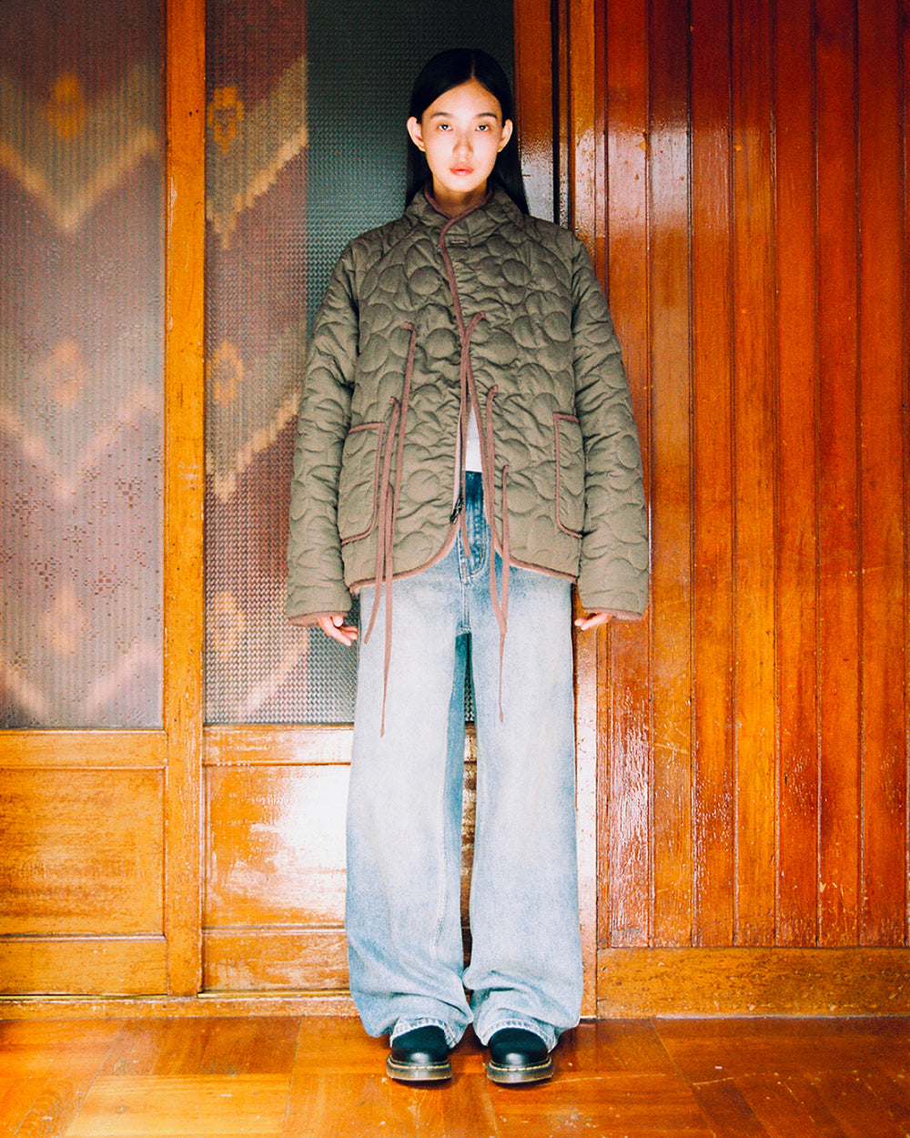 A woman stands indoors wearing the KHAKI REVERSIBLE FLOWER QUILTED JEOGORI FLEECE by Sundae School, paired with wide-leg jeans and black shoes. She is posed against a wooden wall with patterned glass panels, looking directly at the camera with a neutral expression.