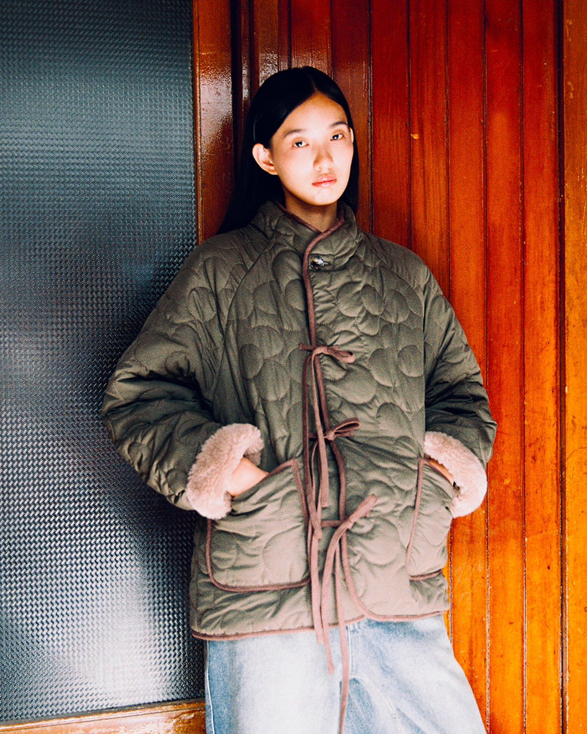 A person wearing the Sundae School KHAKI REVERSIBLE FLOWER QUILTED JEOGORI FLEECE, featuring a hanbok-inspired design and fur-trimmed cuffs, stands against a wooden wall with their hands in the pockets, looking at the camera.