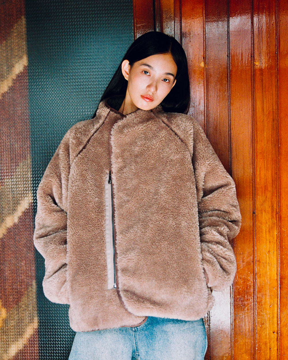 A person with long dark hair stands in front of a wooden wall, calmly looking at the camera with their hands in their pockets, wearing blue jeans and the Sundae School KHAKI REVERSIBLE FLOWER QUILTED JEOGORI FLEECE.