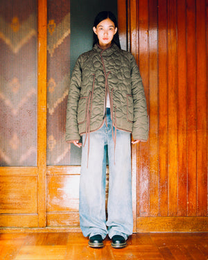 A woman stands indoors wearing the KHAKI REVERSIBLE FLOWER QUILTED JEOGORI FLEECE by Sundae School, paired with wide-leg jeans and black shoes. She is posed against a wooden wall with patterned glass panels, looking directly at the camera with a neutral expression.