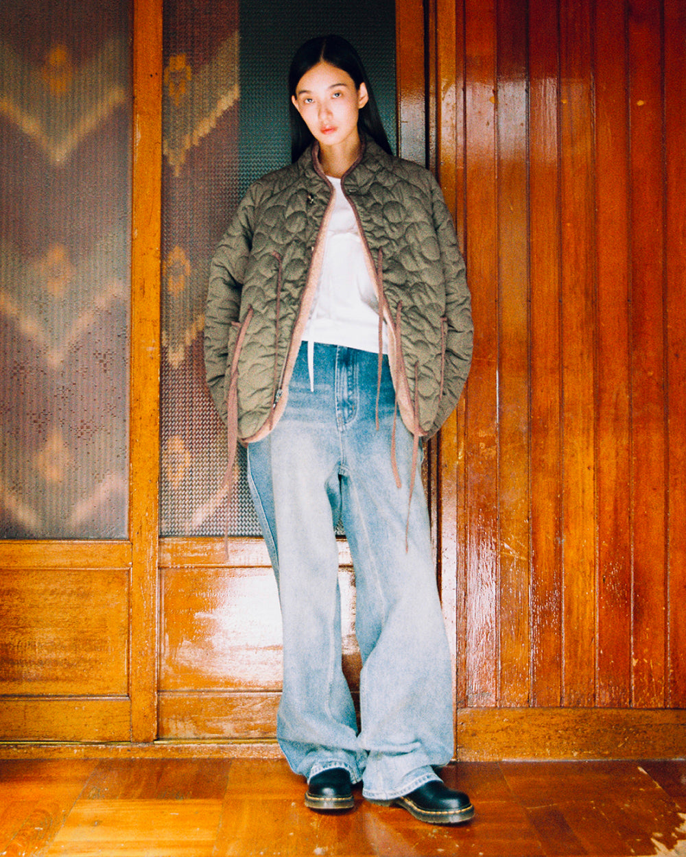 A person stands indoors against wooden doors, wearing the Sundae School KHAKI REVERSIBLE FLOWER QUILTED JEOGORI FLEECE layered over a white top, wide-leg blue jeans, and black shoes, looking at the camera with hands in pockets.