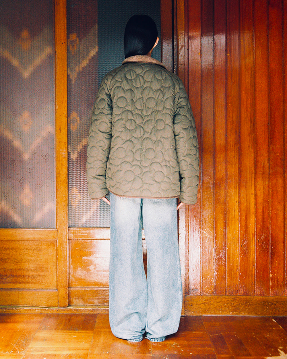 A person with long dark hair stands facing a wooden wall, wearing Sundae School’s Khaki Reversible Flower Quilted Jeogori Fleece and loose-fitting light blue jeans.