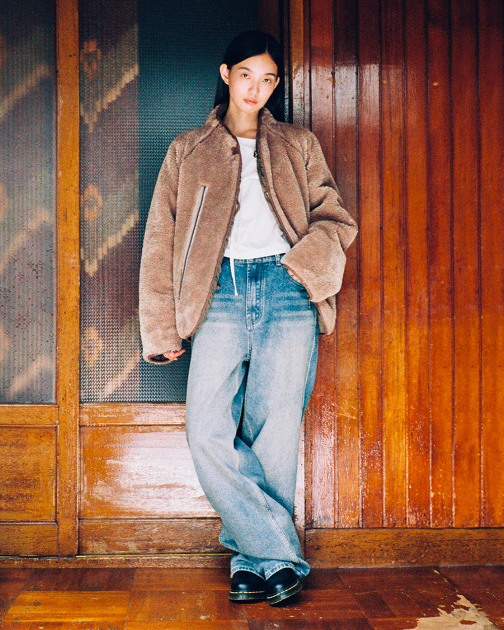 A person stands indoors against a wooden wall, wearing the Sundae School KHAKI REVERSIBLE FLOWER QUILTED JEOGORI FLEECE over a white shirt, paired with loose blue jeans and black shoes. They have straight dark hair and are looking at the camera with a neutral expression.
