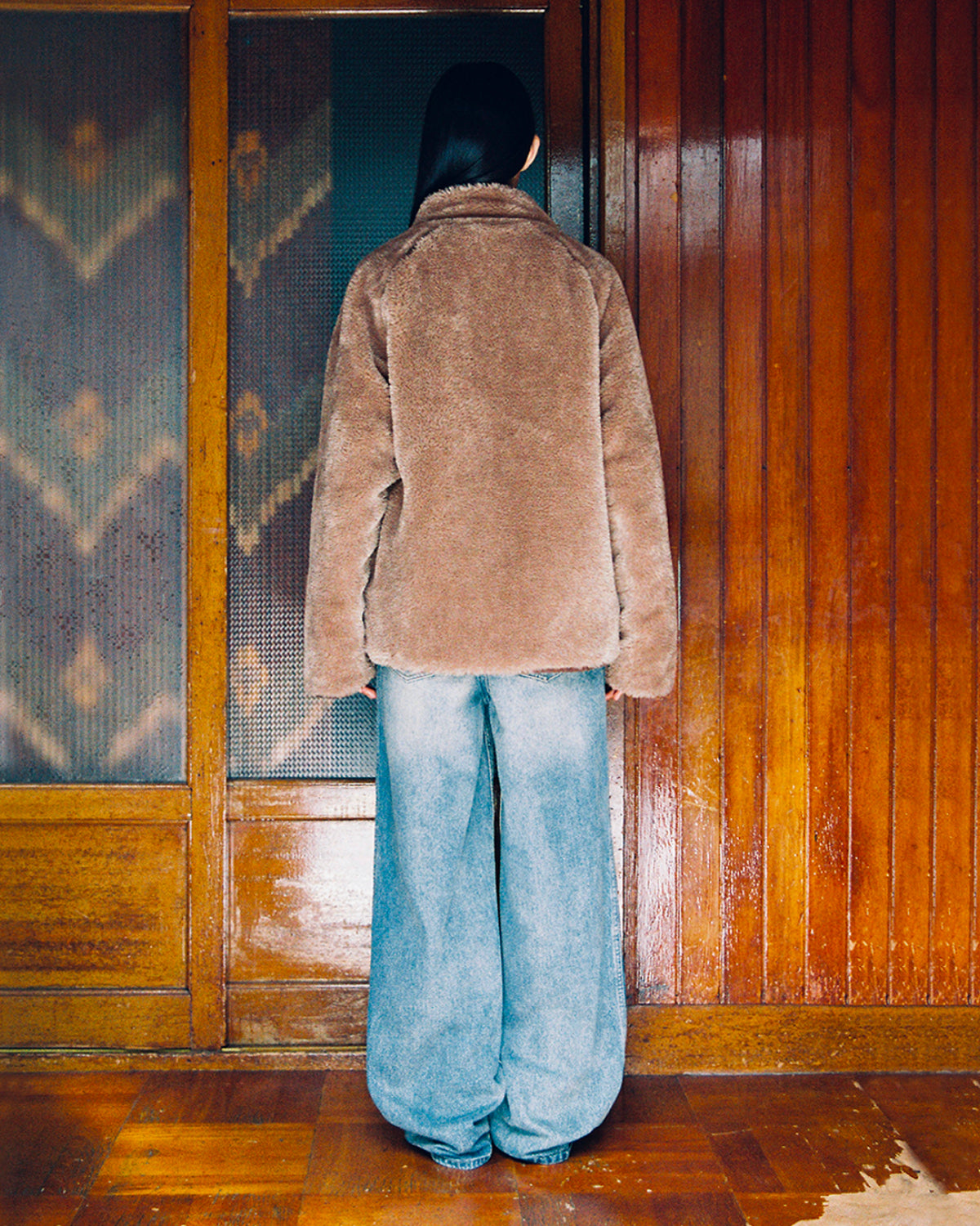 A person with long dark hair stands facing a wooden wall, wearing the Sundae School KHAKI REVERSIBLE FLOWER QUILTED JEOGORI FLEECE and wide-legged blue jeans. Their face is not visible.