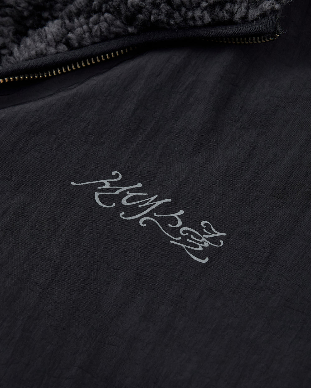 Close-up of the BLACK REVERSIBLE IREZUMI FLEECE by Sundae School, featuring a black textured fabric and a zipper. The jacket showcases gray stylized script that reads “HUMAN,” with a jacquard fleece lining visible at the top edge for added warmth and comfort.