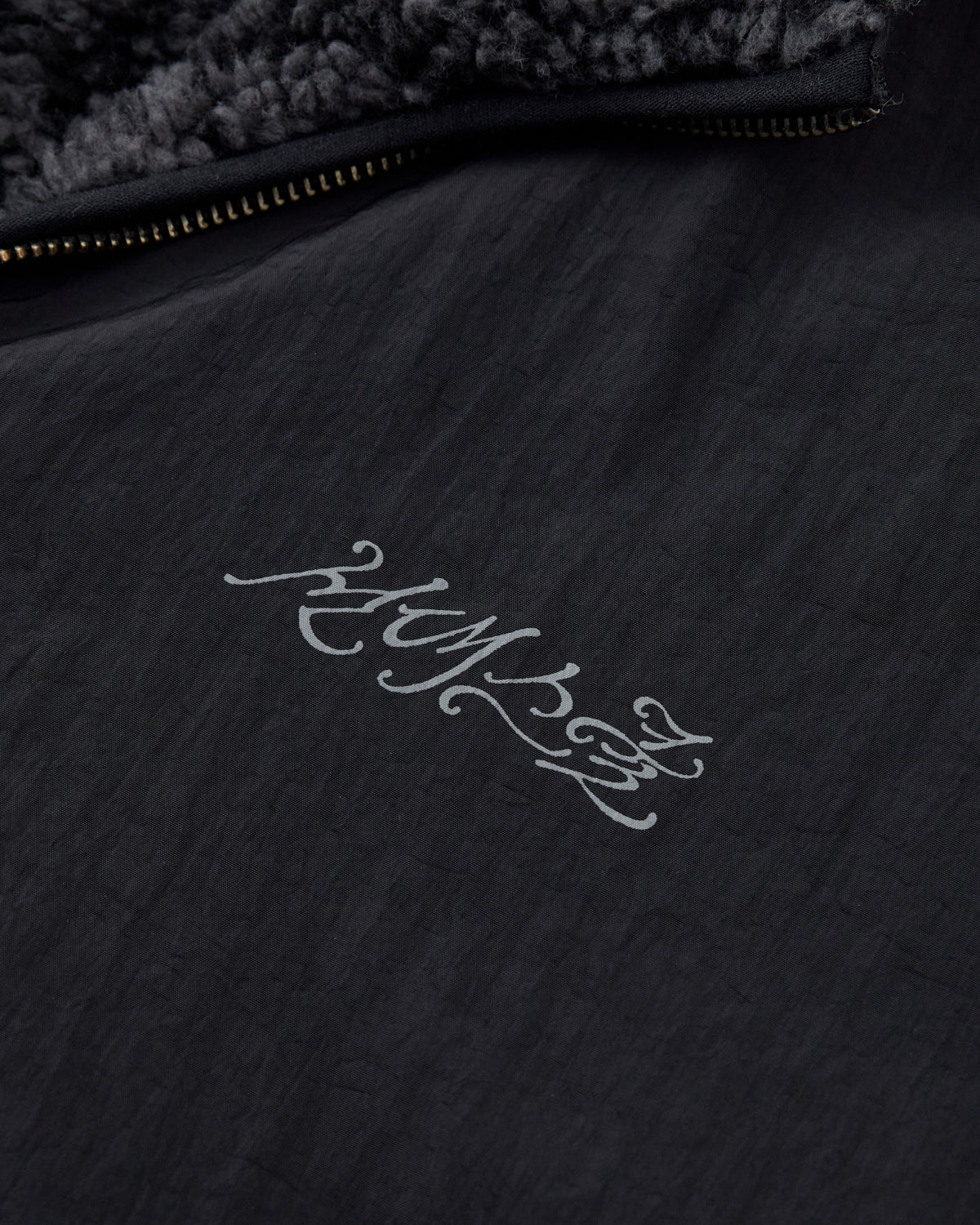 Close-up of the BLACK REVERSIBLE IREZUMI FLEECE by Sundae School, featuring a black textured fabric and a zipper. The jacket showcases gray stylized script that reads “HUMAN,” with a jacquard fleece lining visible at the top edge for added warmth and comfort.