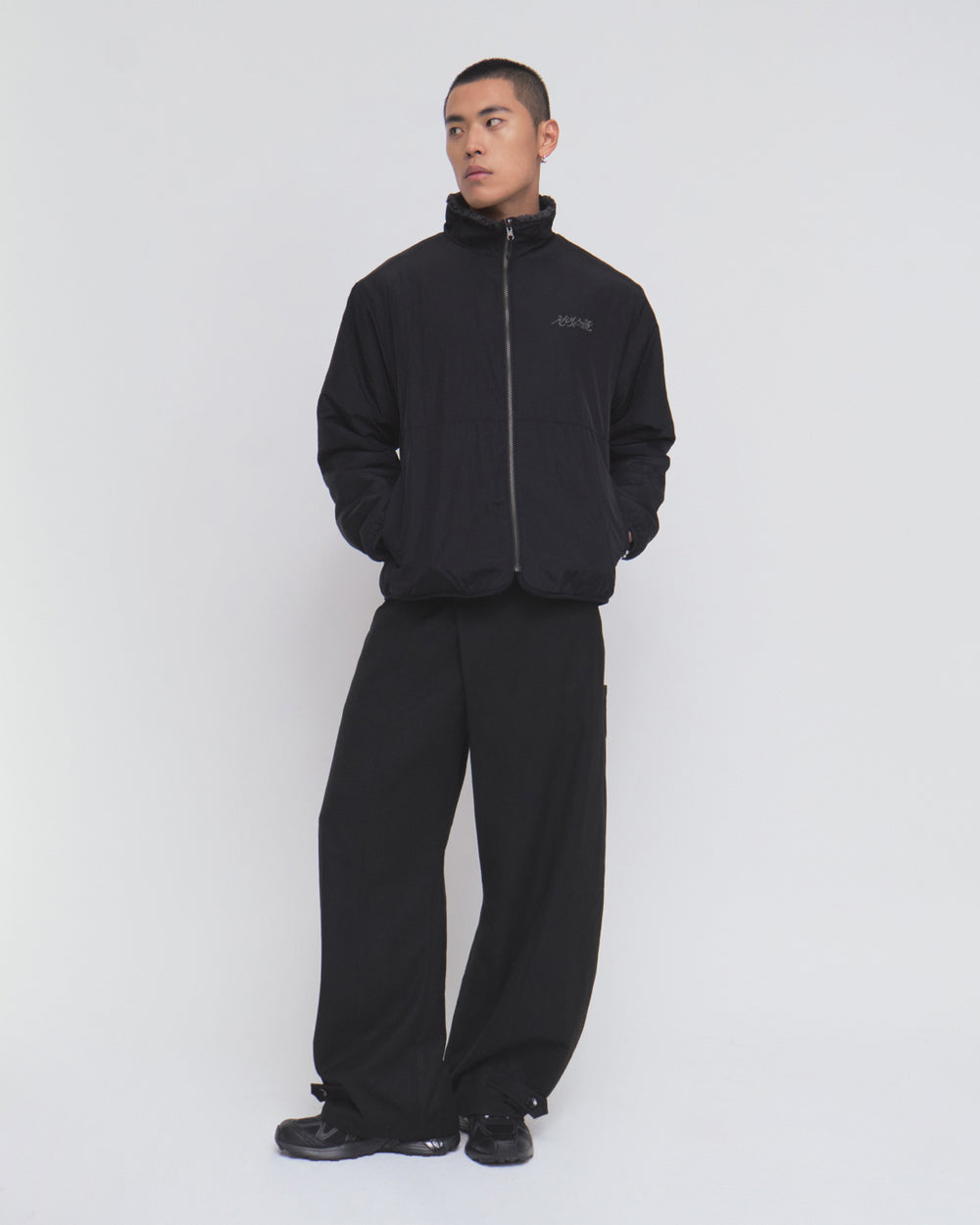 A person stands against a plain white background, wearing the Sundae School BLACK REVERSIBLE IREZUMI FLEECE jacket with their hands in the pockets. The jacket showcases an intricate tiger and dragon graphic. They pair it with loose black pants and black sneakers, gazing slightly to the side.
