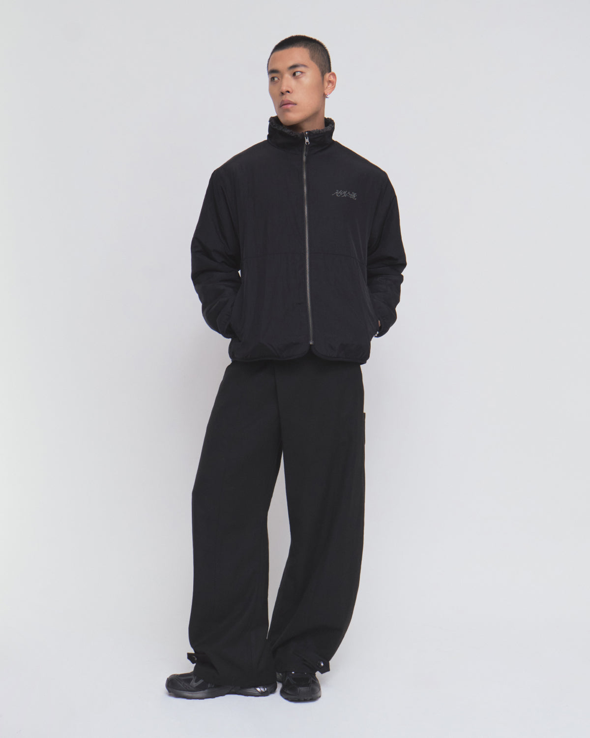 A person stands against a plain white background, wearing the Sundae School BLACK REVERSIBLE IREZUMI FLEECE jacket with their hands in the pockets. The jacket showcases an intricate tiger and dragon graphic. They pair it with loose black pants and black sneakers, gazing slightly to the side.