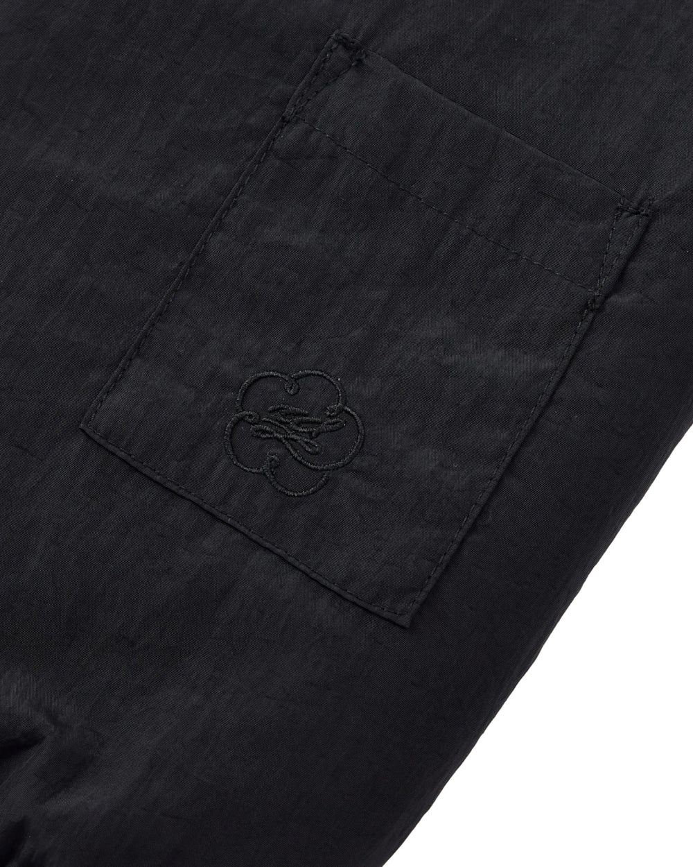 Close-up of a black fabric pocket on the Sundae School BLACK REVERSIBLE IREZUMI FLEECE, featuring a subtle embroidered logo with a stylized flower or clover design. The textured fabric resembles jacquard fleece and appears slightly wrinkled.