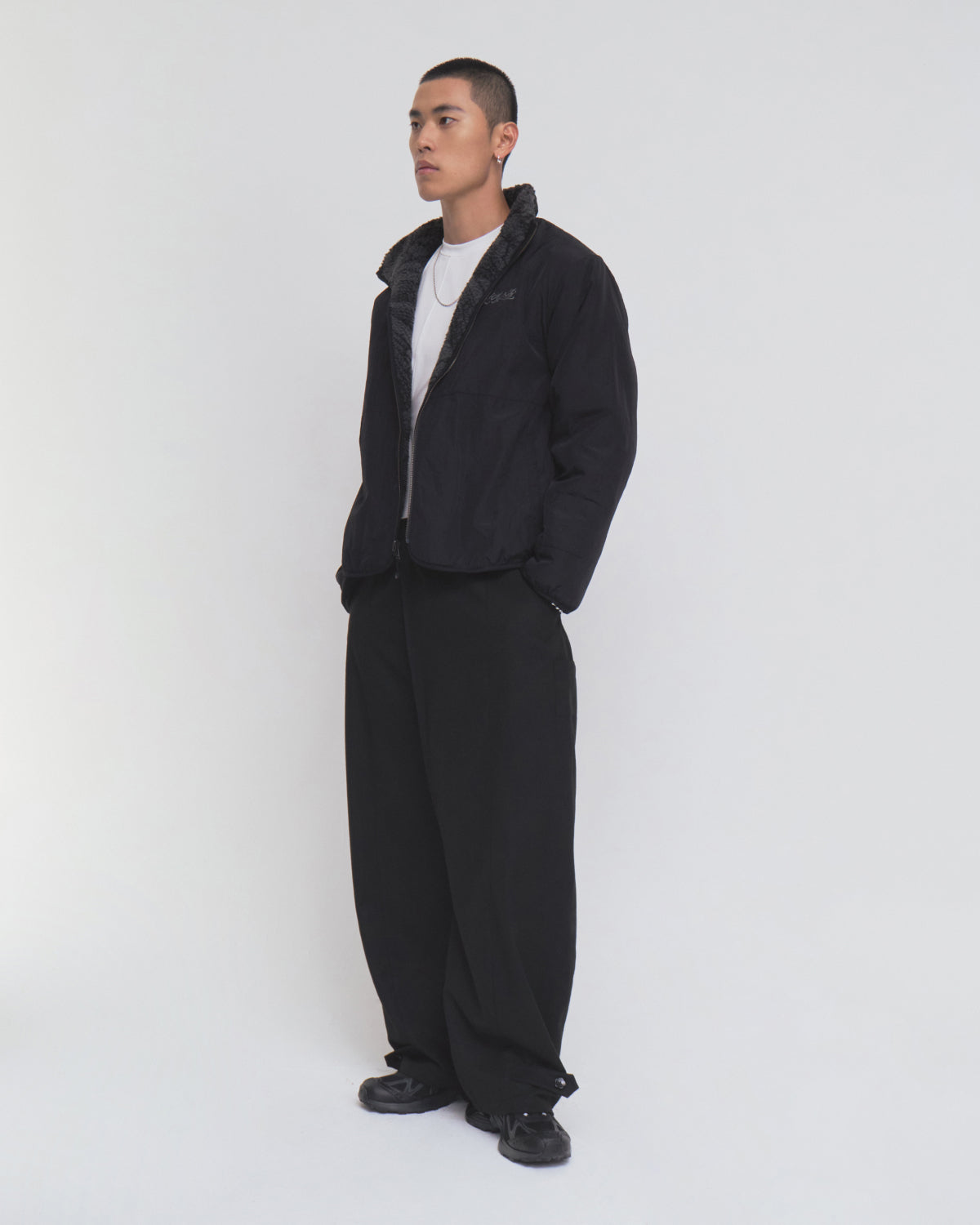A person stands against a plain white background, wearing the Sundae School BLACK REVERSIBLE IREZUMI FLEECE featuring a subtle tiger and dragon graphic, paired with wide black pants, black shoes, and a white T-shirt. Their hands are tucked into the jacket pockets as they look slightly to the side.