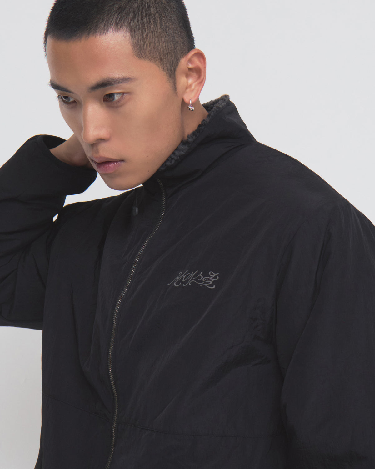 A person with short hair, wearing the Sundae School BLACK REVERSIBLE IREZUMI FLEECE featuring a small embroidered logo, looks to the side while touching the back of their neck.