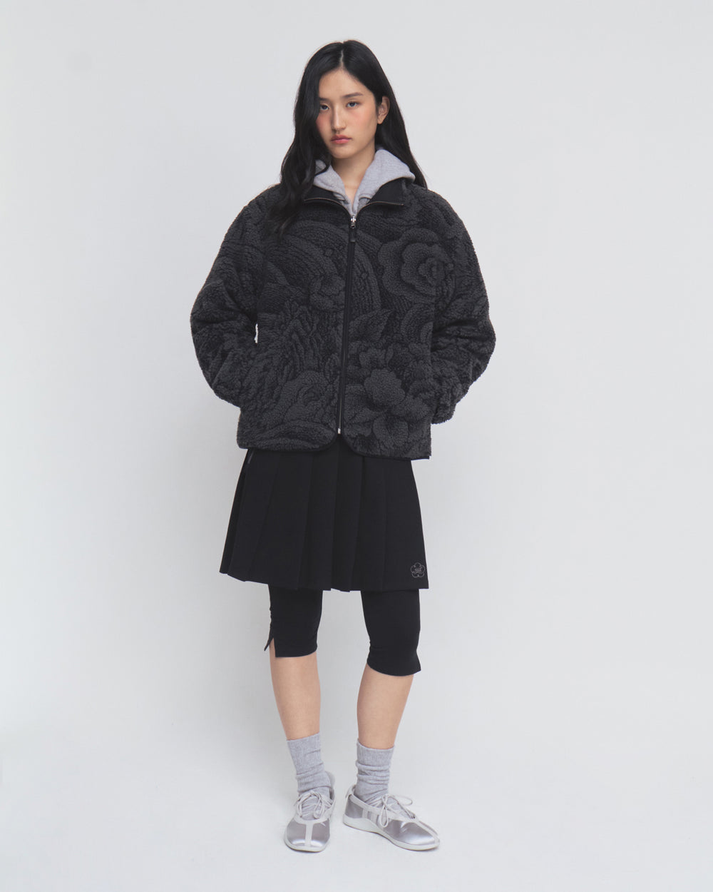 A person stands against a white background wearing the Sundae School BLACK REVERSIBLE IREZUMI FLEECE jacket, styled with a pleated black skirt layered over black leggings, gray socks, and gray sandals.