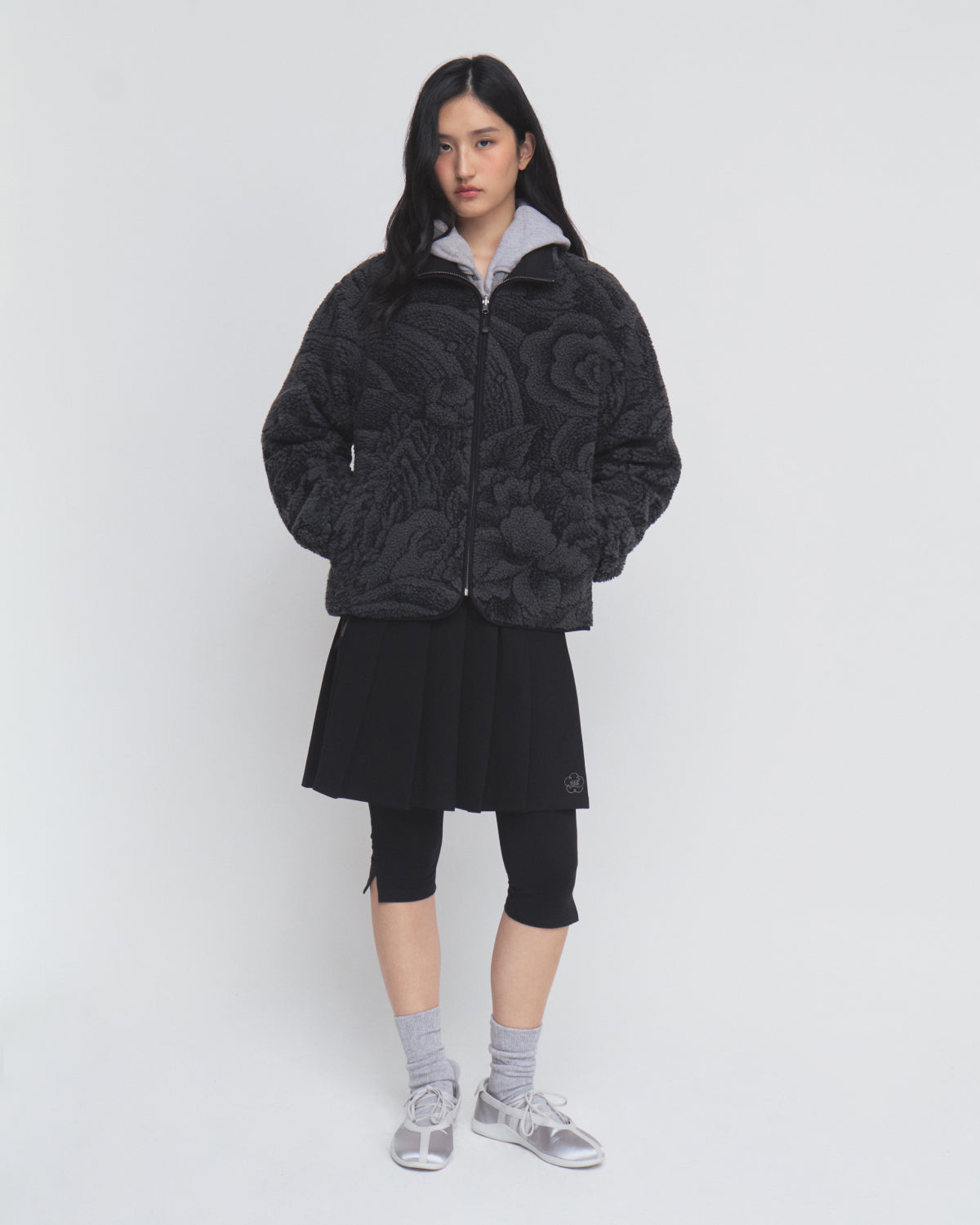 A person stands against a white background wearing the Sundae School BLACK REVERSIBLE IREZUMI FLEECE jacket, styled with a pleated black skirt layered over black leggings, gray socks, and gray sandals.