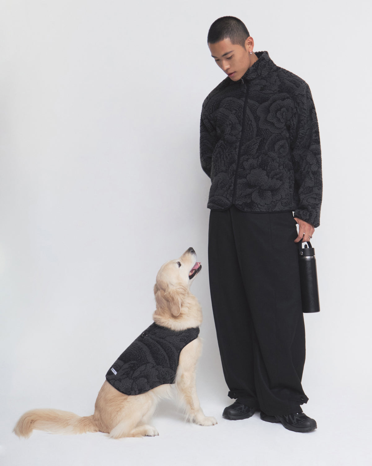 A man dressed in Sundae School’s BLACK REVERSIBLE IREZUMI FLEECE holds a water bottle and looks down at a golden retriever, who is also wearing the matching BLACK REVERSIBLE IREZUMI FLEECE. Both are set against a plain white background.