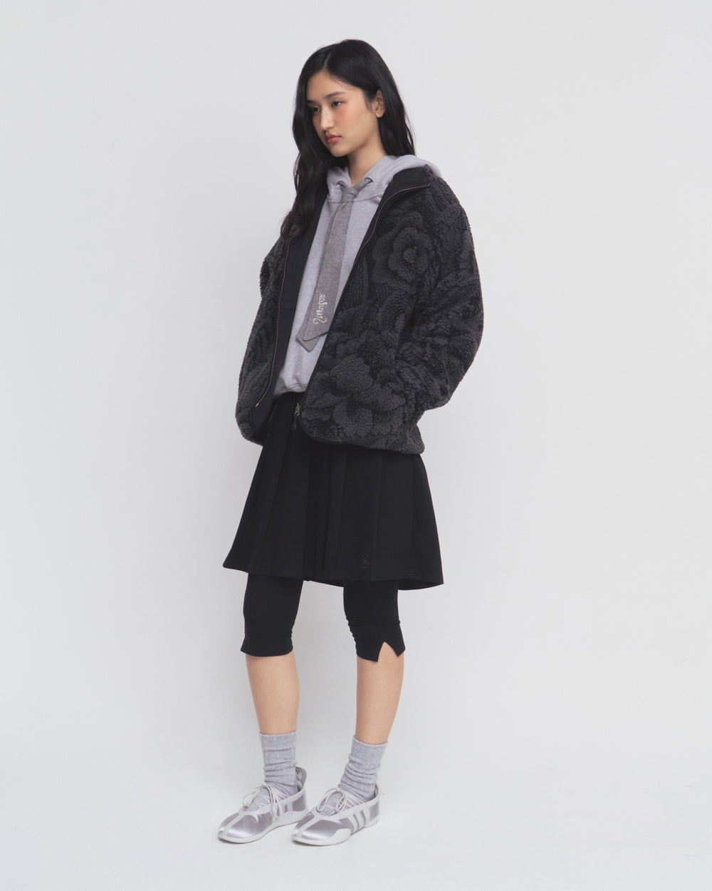 A woman stands against a white background wearing the Sundae School BLACK REVERSIBLE IREZUMI FLEECE, styled with a gray hoodie, black skirt, black leggings, gray socks, and gray sneakers. She has long dark hair and looks slightly downward.