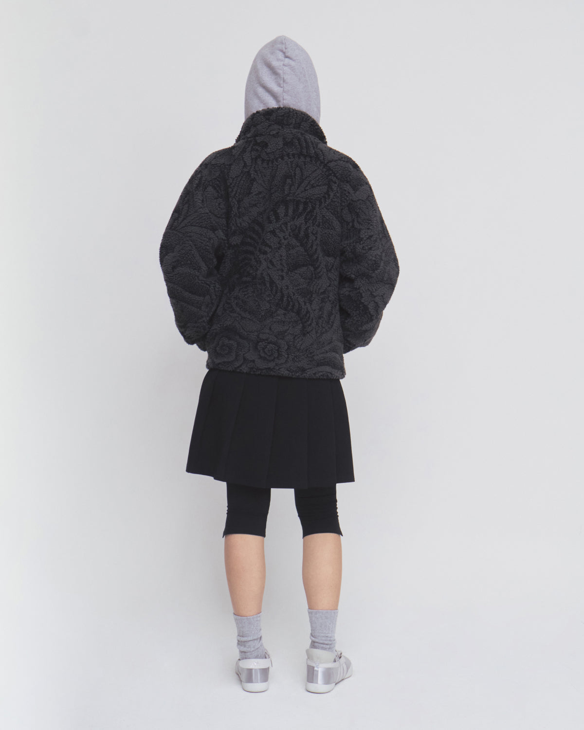 A person stands facing away, wearing a gray hoodie, Sundae School’s BLACK REVERSIBLE IREZUMI FLEECE—a textured dark jacquard jacket—paired with a black skirt over black leggings, gray socks, and translucent shoes against a plain light background.