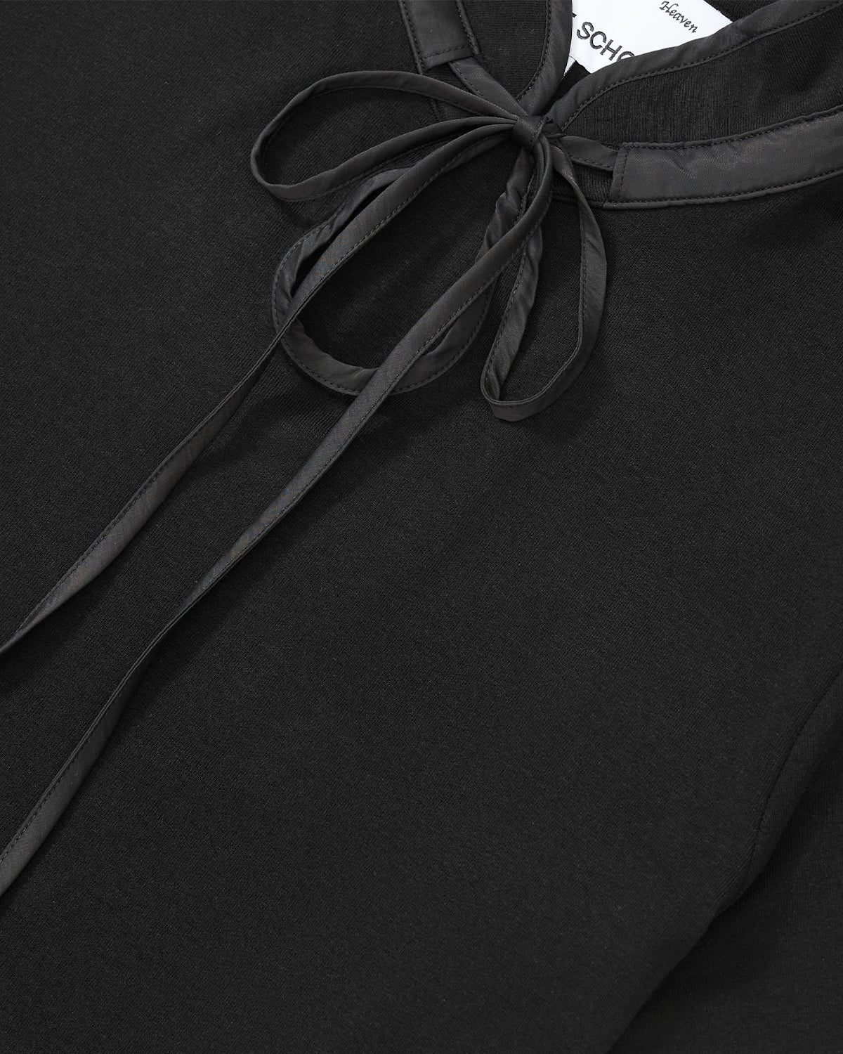 Close-up of the Sundae School BLACK RIBBON TIE POLO showing its Manchurian teardrop collar with satin trim and ribbon tie. The smooth black fabric highlights a partially visible white label inside the collar.