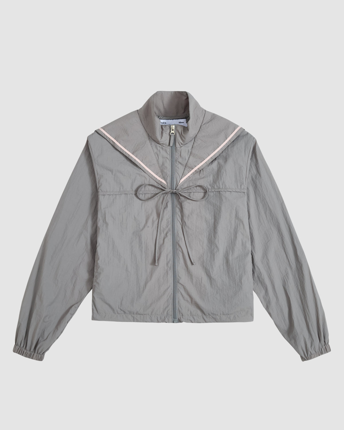 The CHARCOAL SAILOR WINDBREAKER by Sundae School is a light gray cropped jacket with a sailor collar, pink trim, front tie, and long sleeves with elastic cuffs, shown against a plain white background.